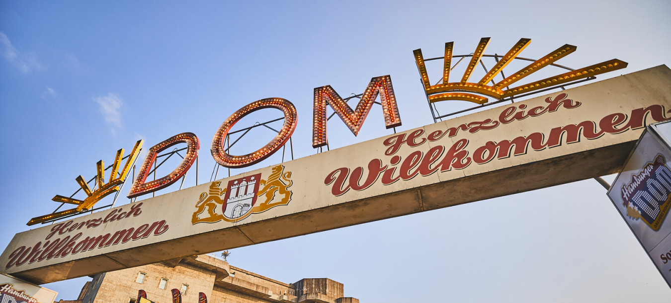 Entrance archway to Hamburg DOM fair with Herzlich Willkommen and decorative lights, festival stalls, and blue sky in the background.