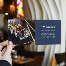 Eine Hand hält einen Geschenkgutschein für das EAST Hotel mit Sushi und dem Text GESCHENK GUTSCHEIN in einem modernen Restaurantambiente.