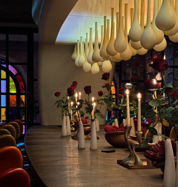 Long wooden table with lit candles in white holders and red roses in vases under a ceiling with hanging teardrop-shaped lamps.