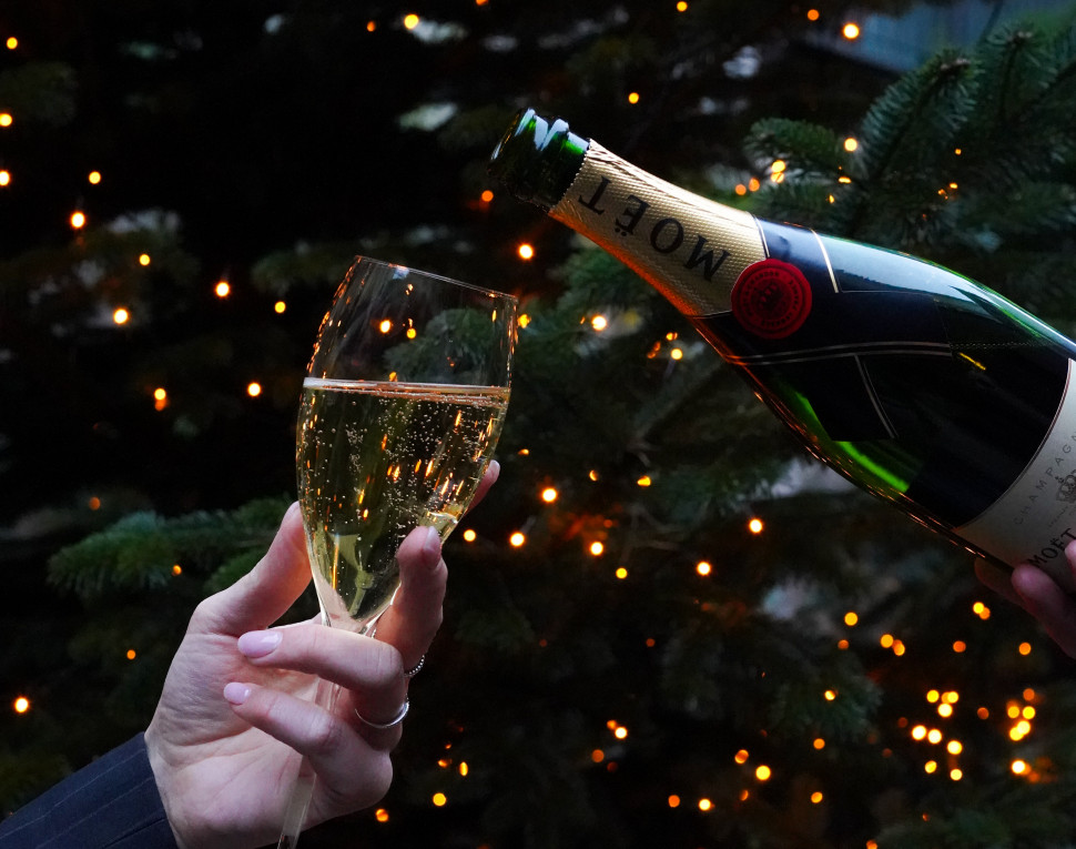 Eine Hand gießt prickelnden Champagner aus einer Flasche in ein Glas vor dem Hintergrund eines Weihnachtsbaums, der mit warmen, leuchtenden Lichterketten geschmückt ist. Festlich und feierlich.