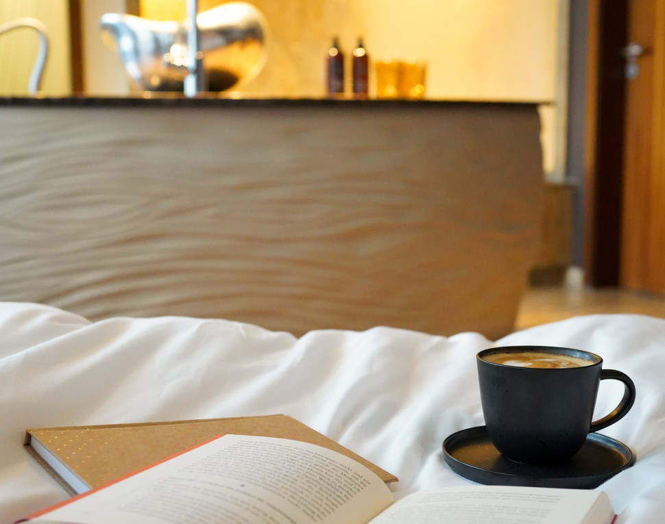 Eine gemütliche Szene mit einem aufgeschlagenen Buch und einem Notizbuch auf einem Bett, dazu eine dampfende Tasse Kaffee. Im Hintergrund ist ein modernes Badezimmer zu sehen, das eine entspannte Atmosphäre schafft.