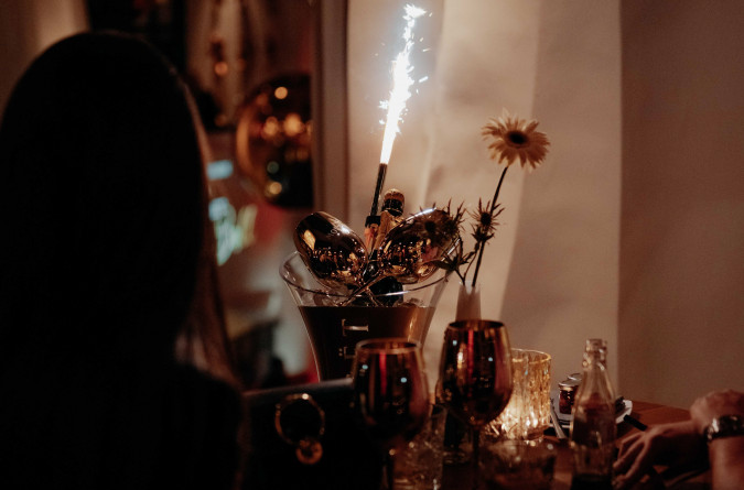 Ein schwach beleuchteter Restauranttisch mit Getränken, einer Blume und einer Sektflasche als Tafelaufsatz. Zwei Personen sitzen, eine davon liegt größtenteils im Schatten.