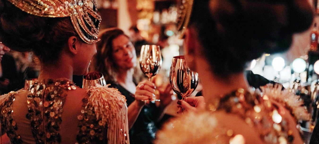 Auf einer festlichen, eleganten Party stoßen Menschen in glamourösen Pailletten-Outfits mit goldenen Weingläsern an.