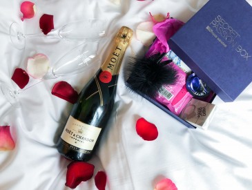 Champagnerflasche, Rosenblätter und Secret Passion Box auf weißer Bettdecke.