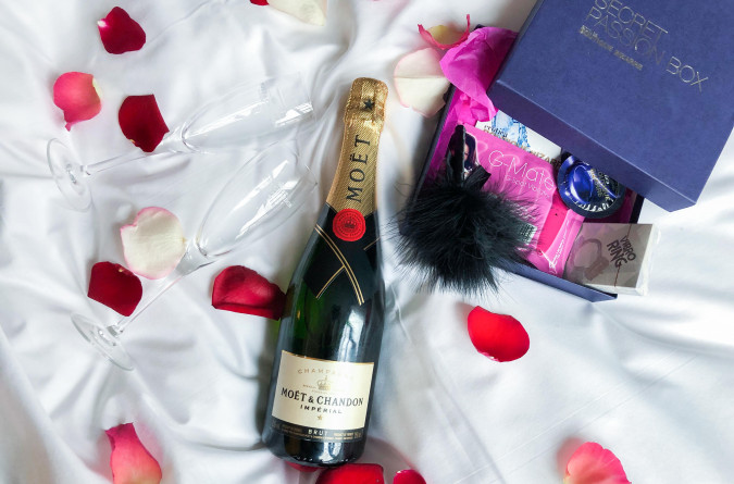 Secret Passion Kit mit Champagnerflasche, Gläsern und Rosenblättern auf dem Bett