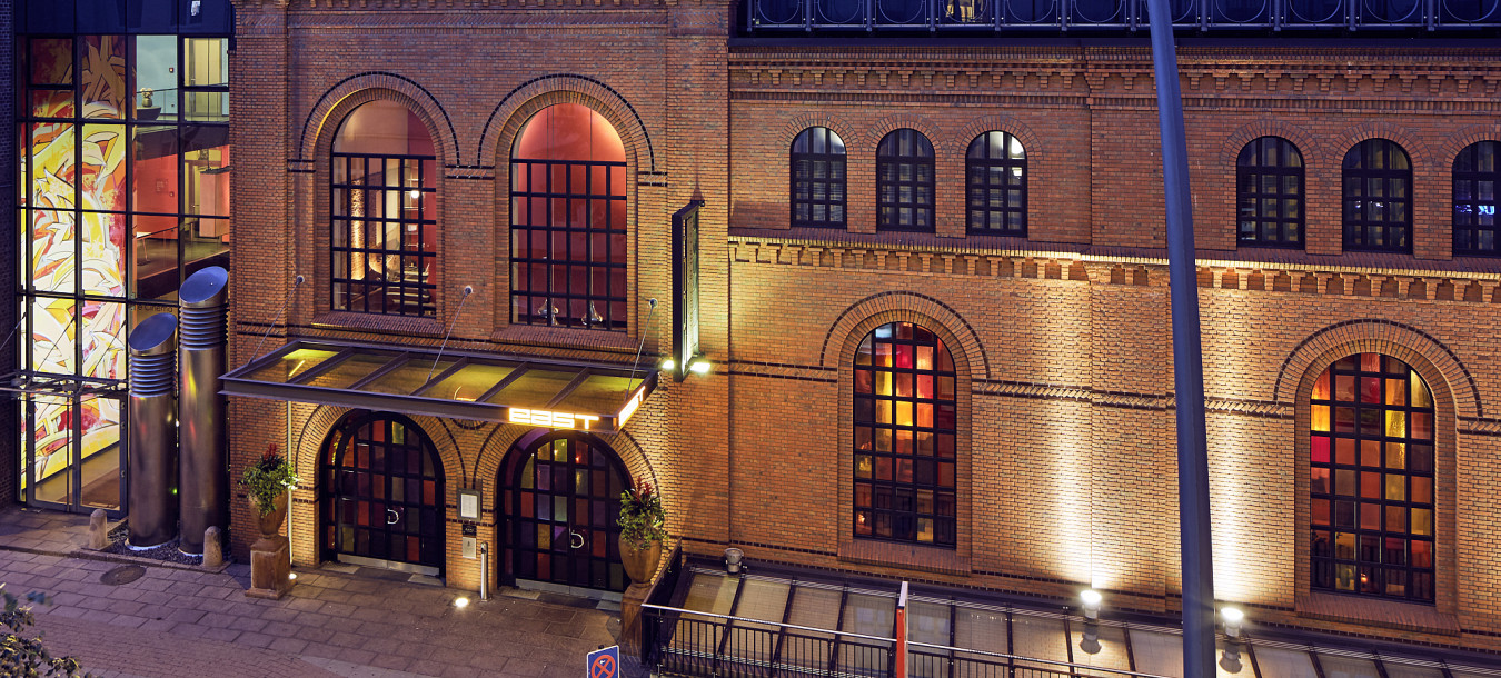 Ein modernes Glasgebäude erhebt sich über einer Backsteinfassade mit Bogenfenstern und wird in der Abenddämmerung auf einer Stadtstraße warm beleuchtet.