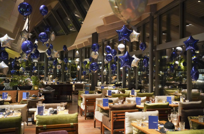 Ein Restaurant, das mit blauen und silbernen Sternballons, gedeckten Esstischen und großen Fenstern mit Blick auf die Lichter der Stadt bei Nacht dekoriert ist.
