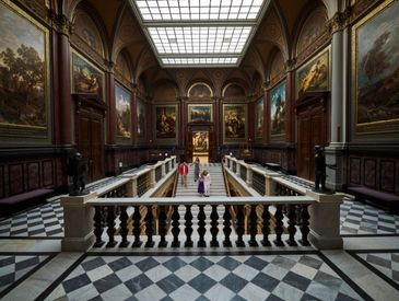 Große Halle mit Schachbrettboden, hohen gewölbten Decken, großen Gemälden, Statuen und Menschen, die eine zentrale Treppe hinuntergehen.