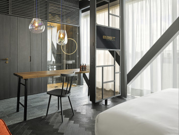 Modern hotel room with a wooden desk, chair, hanging lamps, TV, large windows with transparent curtains, and a visible part of a white bed.