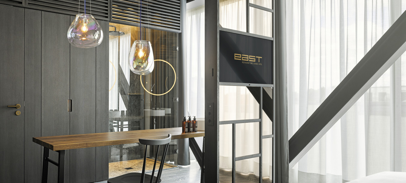 Modern hotel room with a wooden desk, black chair, hanging lights, TV, and large windows with sheer curtains.