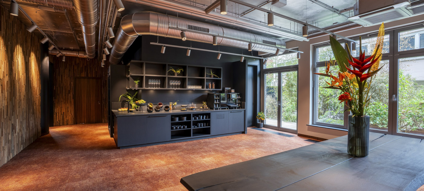 Moderne Büroküche mit freiliegenden Leitungen, eleganten Schränken, großen Fenstern und einem schwarzen Tisch mit einem tropischen Blumenarrangement.