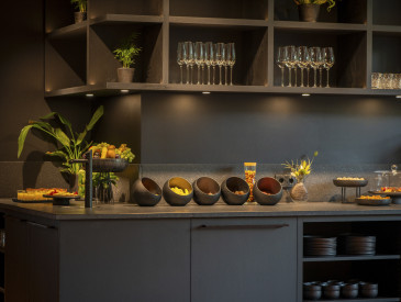 Modern kitchen countertop with snack bowls, glassware, fruit, and decorative plants, with shelves for glasses and plates underneath.