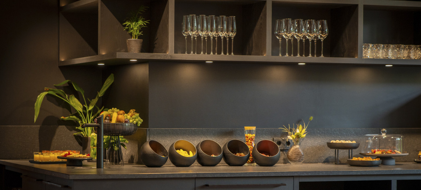 A modern kitchen counter with bowls of snacks, fruit, desserts, glassware, and plants on display shelves and countertops.