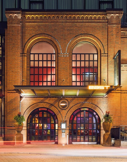 The photo shows the EAST Hotel in Hamburg with its brick facade and large, internally illuminated arched windows. Above the entrance, the lettering 