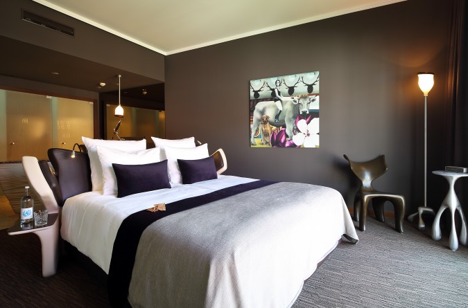 Hotelzimmer mit einem großen Bett mit weißer und grauer Bettwäsche, lilafarbenen Akzentkissen, einem modernen Stuhl und Nachttischen, einer Stehlampe und einem farbenfrohen Kunstwerk an einer dunklen Wand. Auf einem Nachttisch steht eine Wasserflasche.