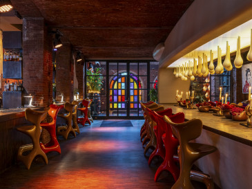 The stylish east lobby bar features curved orange chairs, red velvet seating, exposed brick walls, modern pendant lights, and large stained-glass windows.