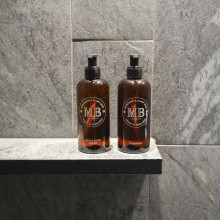 Zwei braune Pumpflaschen mit der Aufschrift „MB“ stehen auf einem Steinregal vor einer grau gekachelten Wand in einer Dusche oder einem Badezimmer.