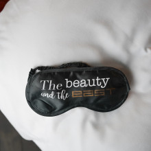 Eine schwarze Schlafmaske mit weißer und goldener Aufschrift „The beauty and the best“ liegt auf einem weißen Kissen. Die Umgebung vermittelt Entspannung.