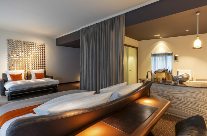 Modernes Zimmer der Kategorie Signature mit großem Bett, Sofa mit Kissen, Trennvorhang, Schreibtisch und stilvollem Badezimmer mit Waschbecken und dekorativer Beleuchtung. Neutrale Töne mit orangefarbenen Akzenten verleihen dem Raum Wärme.