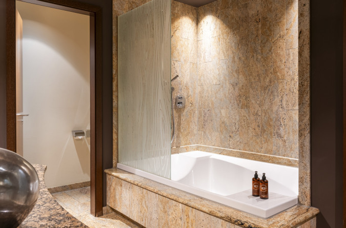 Ein modernes Badezimmer verfügt über eine Badewanne aus beigefarbenem Marmor mit einer dekorativen Duschwand aus Glas, einem an der Wand montierten Duschkopf und zwei Flaschen Toilettenartikeln auf dem Wannenrand; durch eine offene Tür ist eine Toilett