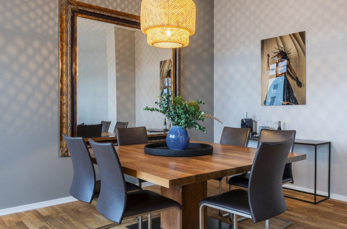 Esszimmer mit einem Holztisch, vier schwarzen Stühlen, einem großen Spiegel an der Wand, einer geflochtenen Hängelampe, einer blauen Vase mit grünem Blattwerk und einem gerahmten Foto. Lichtmuster werden an die Wand geworfen.