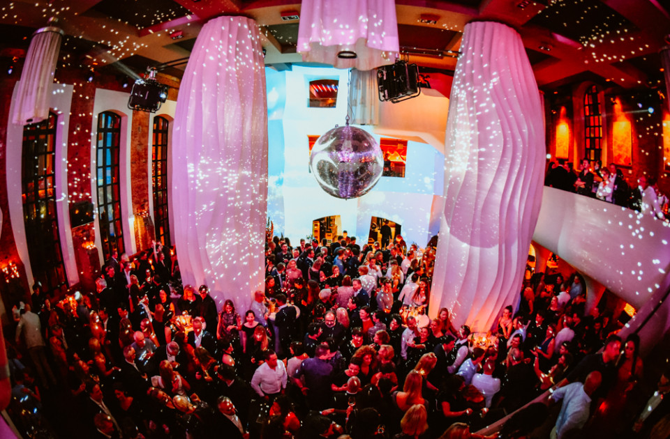 Eine große Menschenmenge genießt eine lebhafte Party in einem glamourösen Veranstaltungsort mit einer Discokugel und bunten Lichtern, die von hohen, weißen Säulen reflektiert werden.