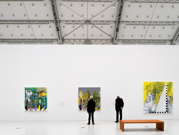 Drei Personen betrachten farbenfrohe abstrakte Gemälde an einer weißen Wand in einer modernen, geräumigen Kunstgalerie mit hoher Decke im Industriestil.