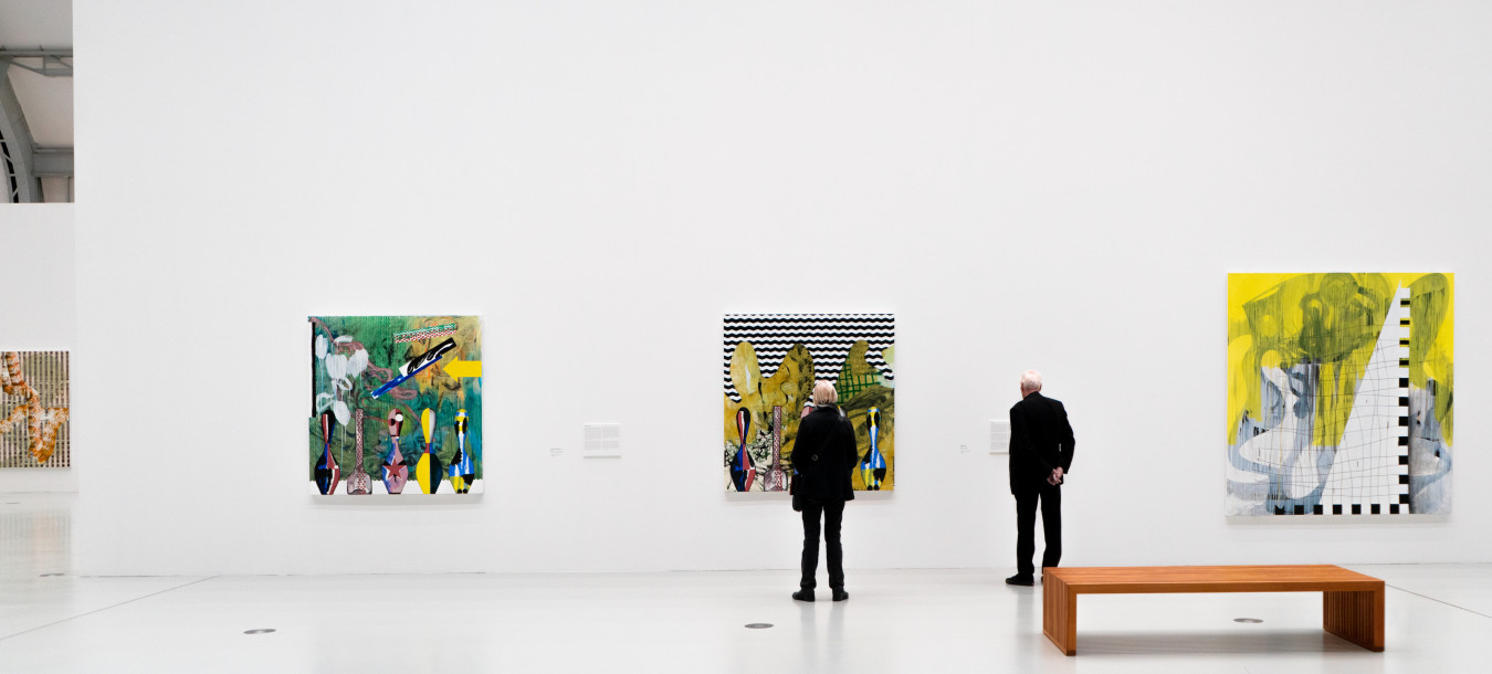 Drei Personen betrachten farbenfrohe abstrakte Gemälde in einer modernen, geräumigen Kunstgalerie mit hoher Decke im Industriestil.