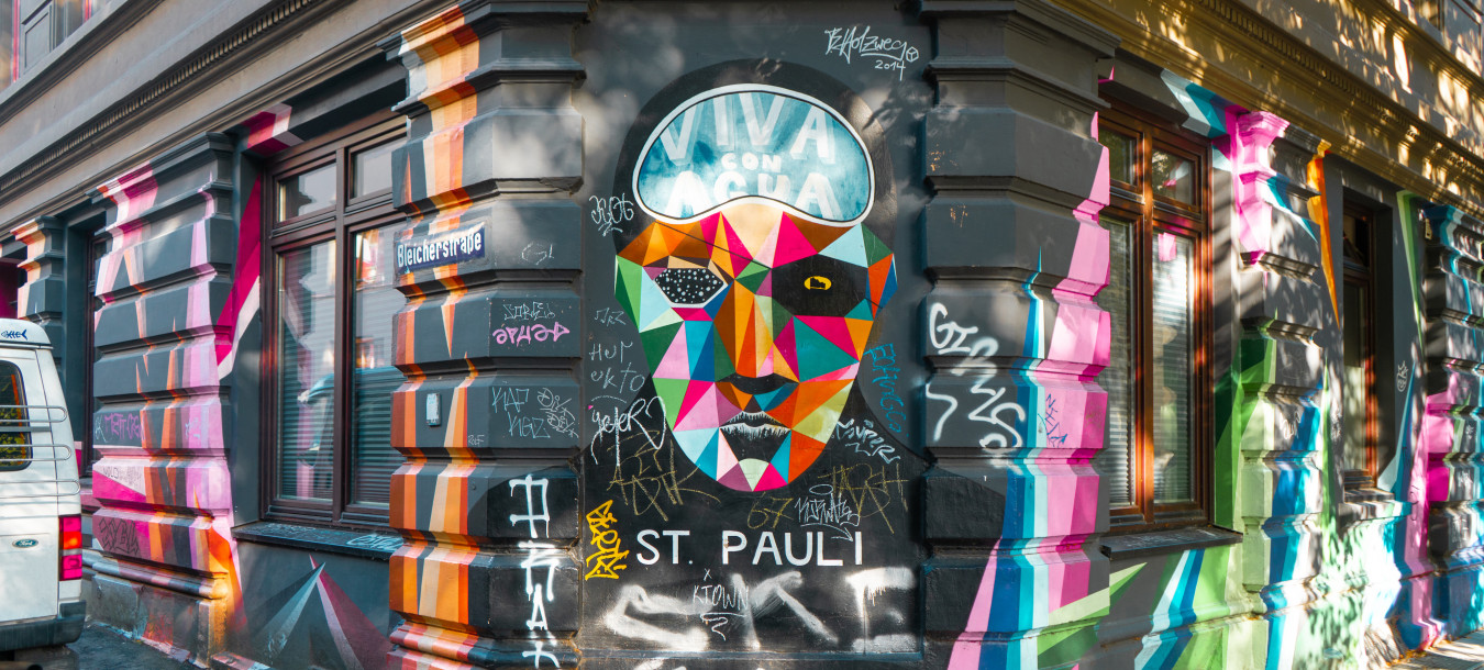 Farbenfrohes geometrisches Gesichtswandbild mit dem Text „Viva con Agua“ an einem Eckgebäude, umgeben von Graffiti und leuchtend gemalten Streifen.