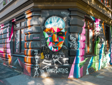 Farbenfrohes Straßenwandbild eines facettierten Gesichts mit einer Viva con Agua-Maske an einem Eckgebäude mit Graffiti und hell aufgemalten Streifen.