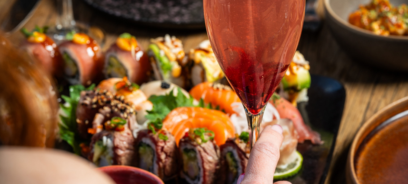 A person holds a pink cocktail while another uses chopsticks to pick up sushi from a platter on a wooden table.