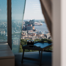Eine Stadtlandschaft mit einem Gebäude am Wasser, das durch ein Fenster gesehen wird, das teilweise von einem Vorhang verdeckt ist, mit einem Tisch im Vordergrund.