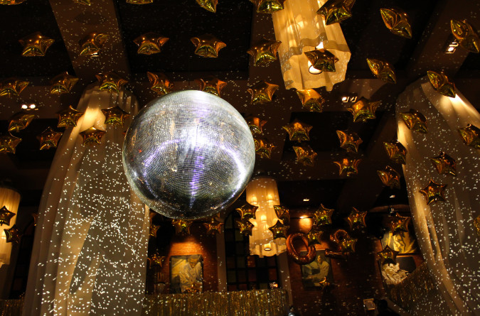 Eine schimmernde Discokugel reflektiert das Licht in einem festlich geschmückten Raum, der mit zahlreichen goldenen sternförmigen Luftballons und eleganten Kronleuchtern dekoriert ist und eine feierliche Atmosphäre schafft.
