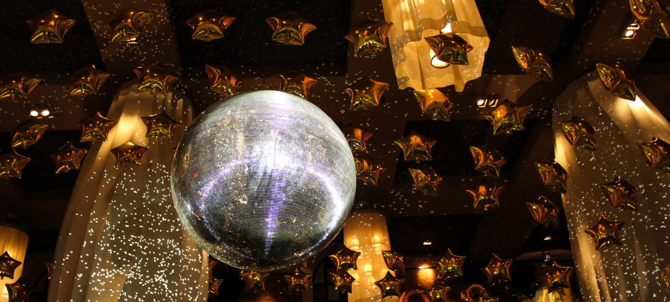 A shimmering disco ball reflects light in a festive room adorned with numerous gold star-shaped balloons and elegant chandeliers, creating a celebratory ambiance.