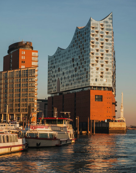 Modern glass building with a wavy roof on the water, with docked boats in the foreground illuminated by golden sunlight.