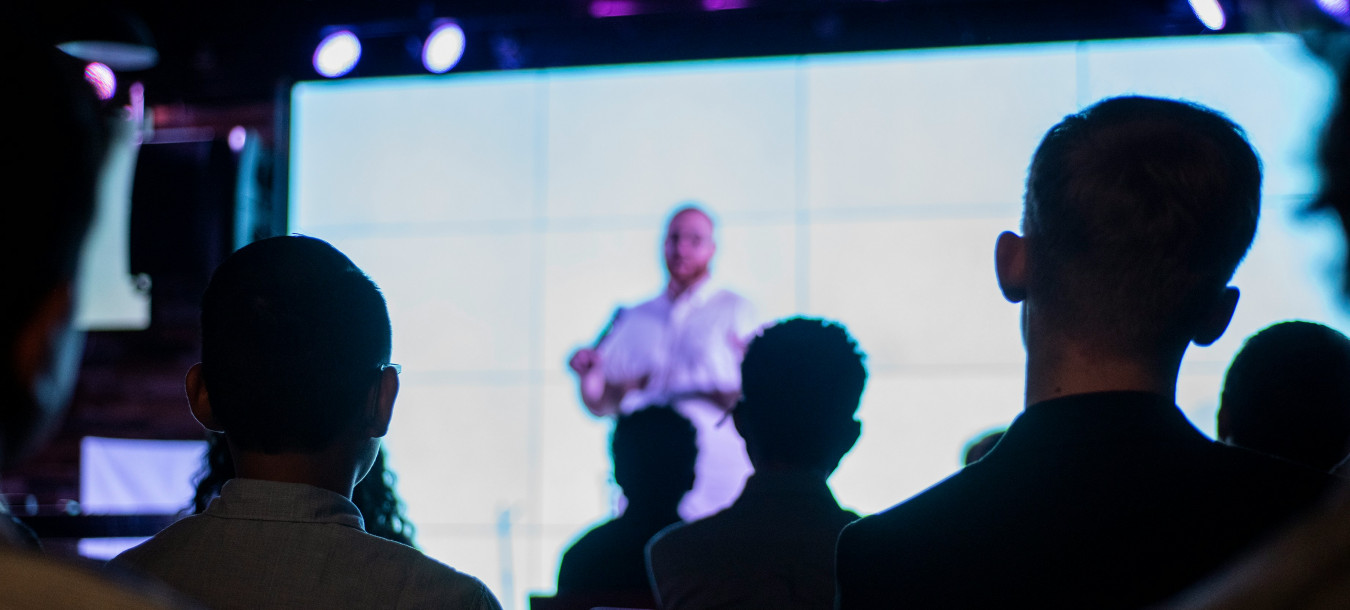 Mehrere Personen sitzen in einem dunklen Raum und schauen auf eine beleuchtete Bühne, auf der eine Person steht und spricht.