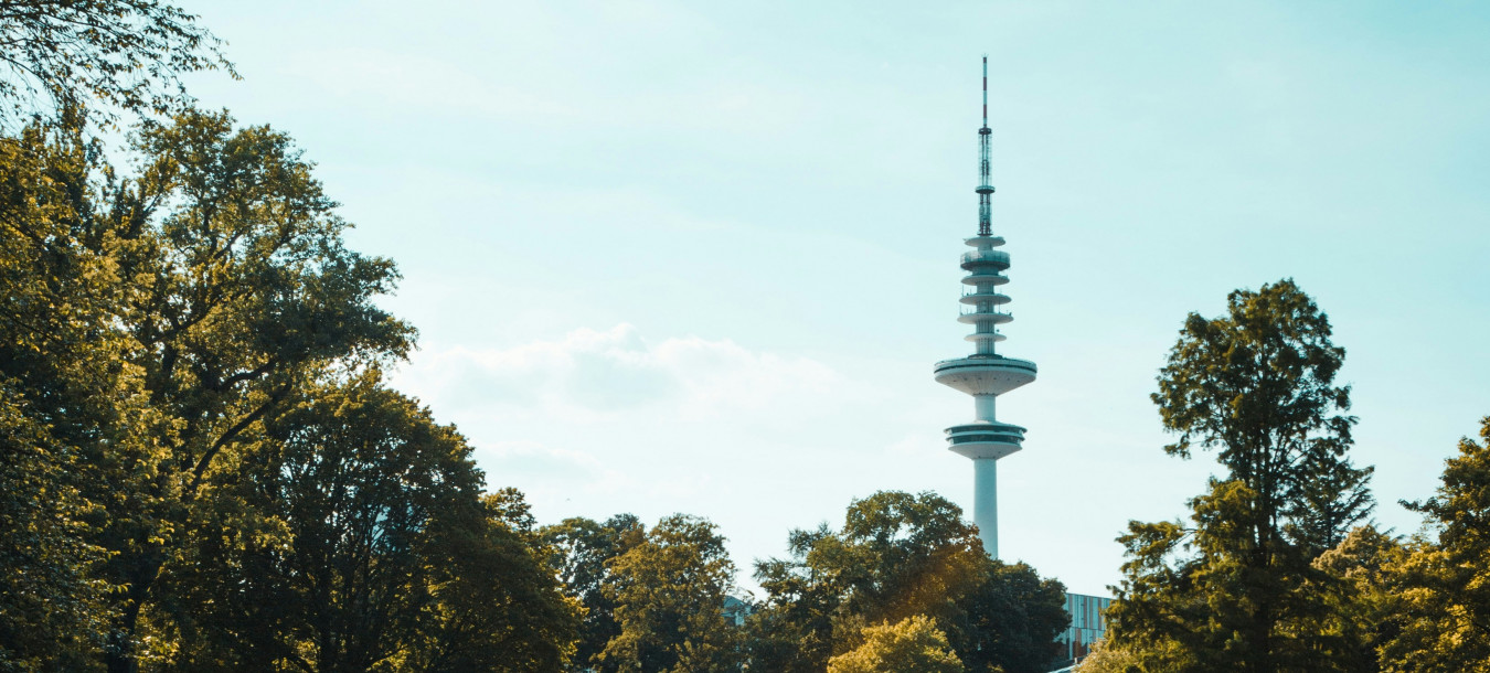 Ein hoher Fernsehturm erhebt sich über Bäumen und einem ruhigen Teich unter einem blauen Himmel mit vereinzelten Wolken in einer Parklandschaft.