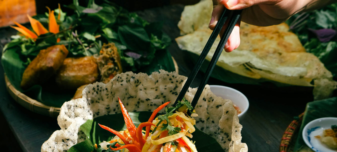 A person uses chopsticks to pick salad from a sesame rice cracker bowl, surrounded by Vietnamese dishes on a wooden table.